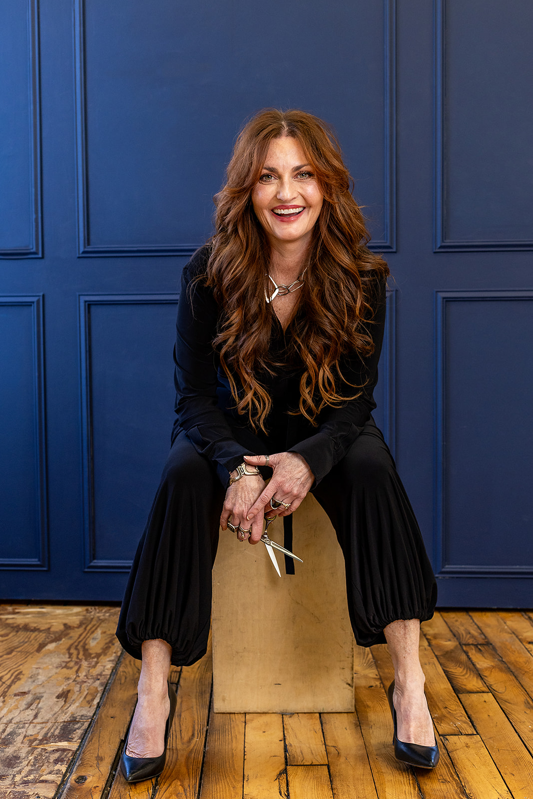 Photo of Julianne holding scissors and sitting, looking at the camera with an open mouth smile.